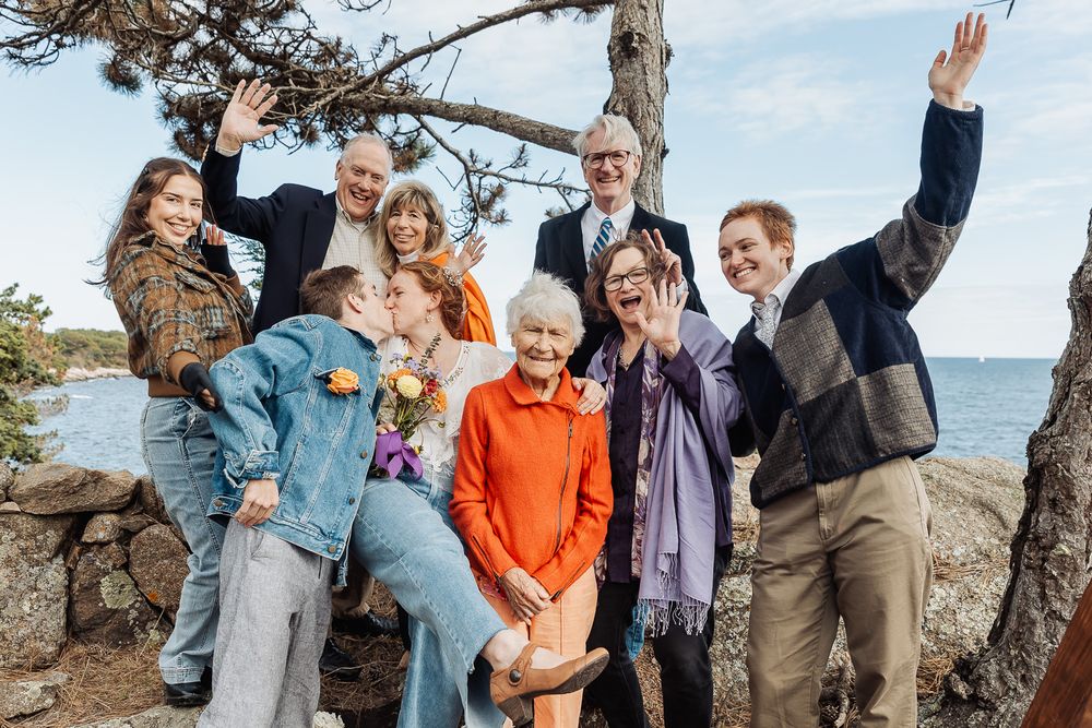 A joyful multigenerational family group celebrates outdoors near rocky coastline, waving and smiling together.