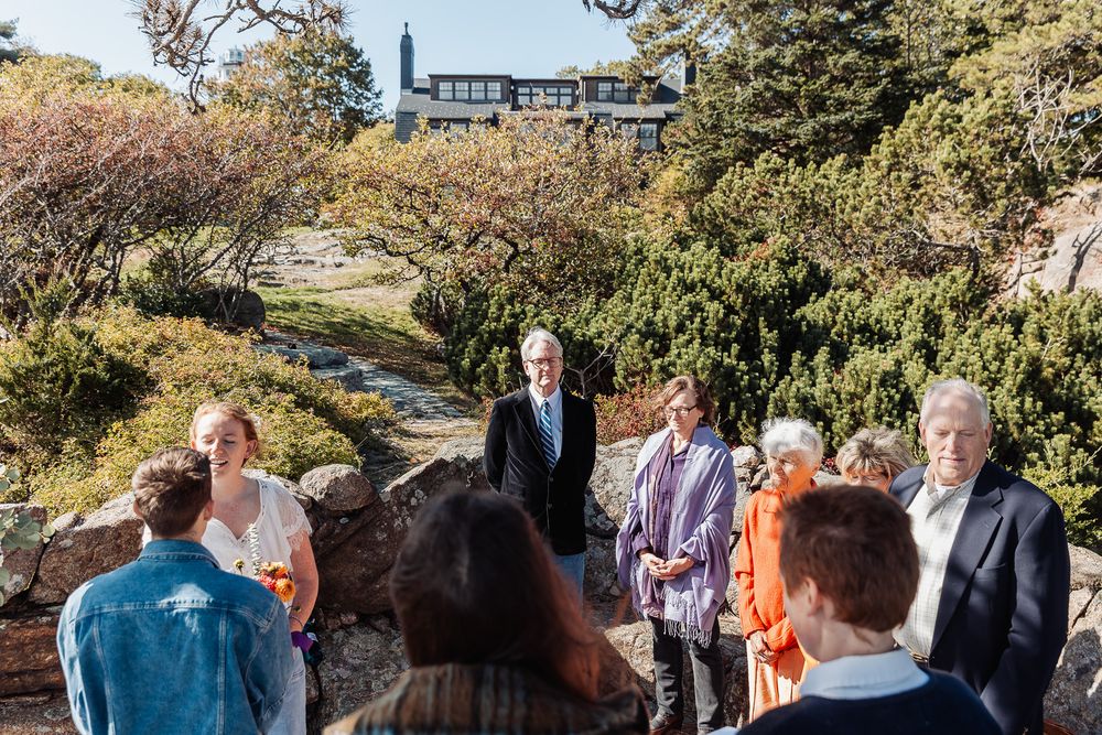Group of people gathered in a scenic rocky garden with lush shrubs and a building in the background on a sunny day.