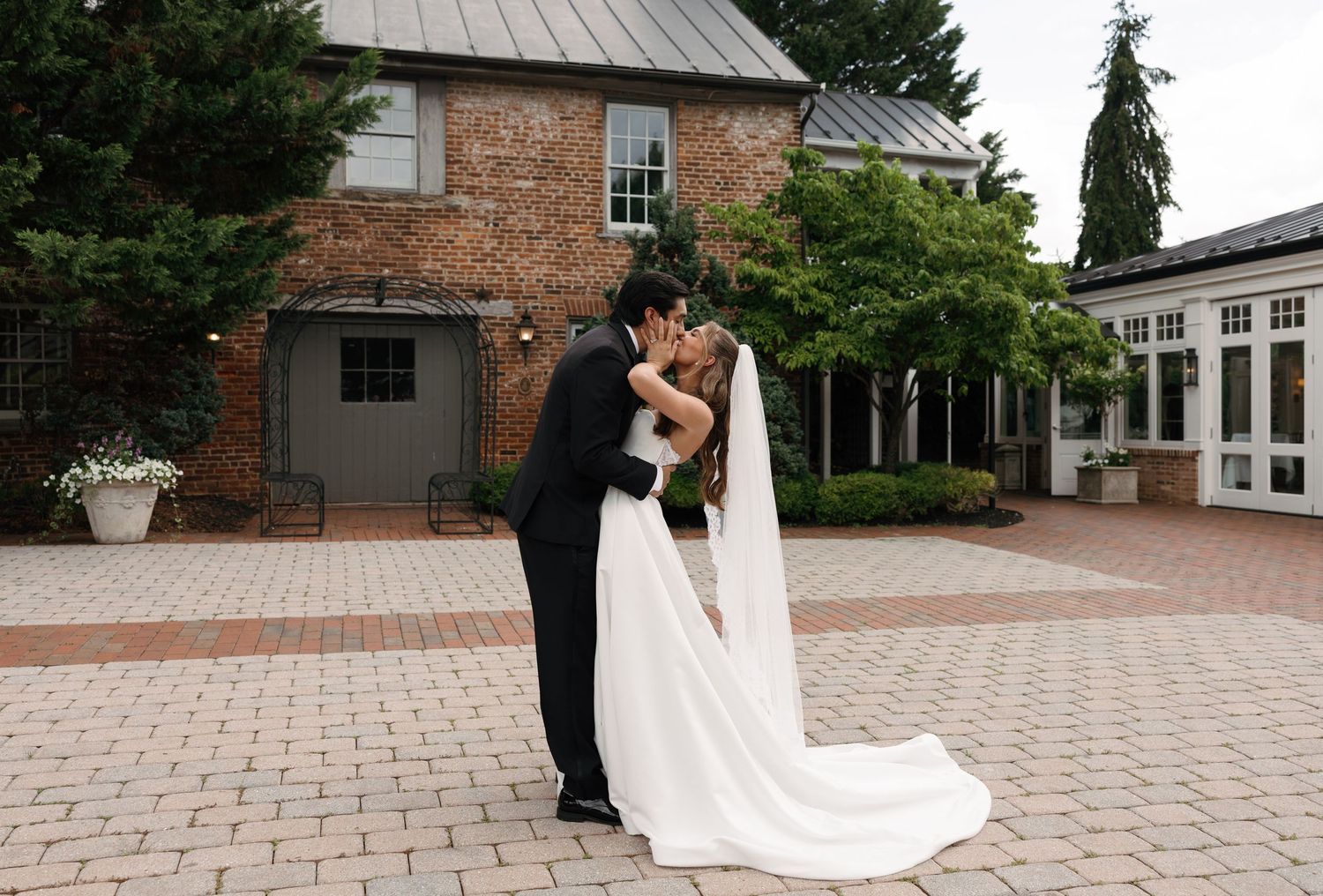 Bride and groom share a romantic kiss in a brick courtyard outside an elegant wedding venue surrounded by lush greenery.