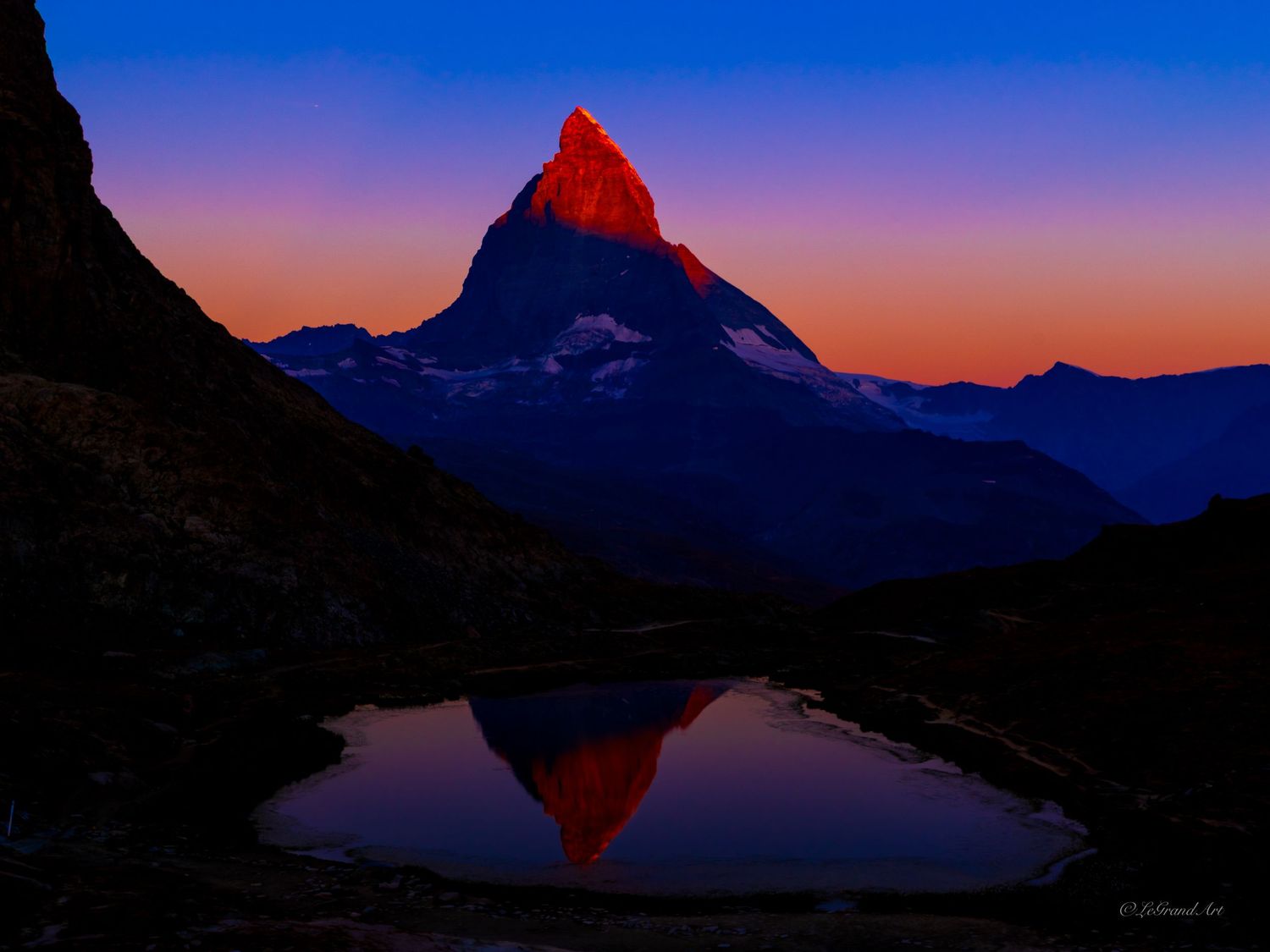 The Matterhorn peak glows red at sunrise, reflected in a calm alpine lake under a vivid blue and orange sky.