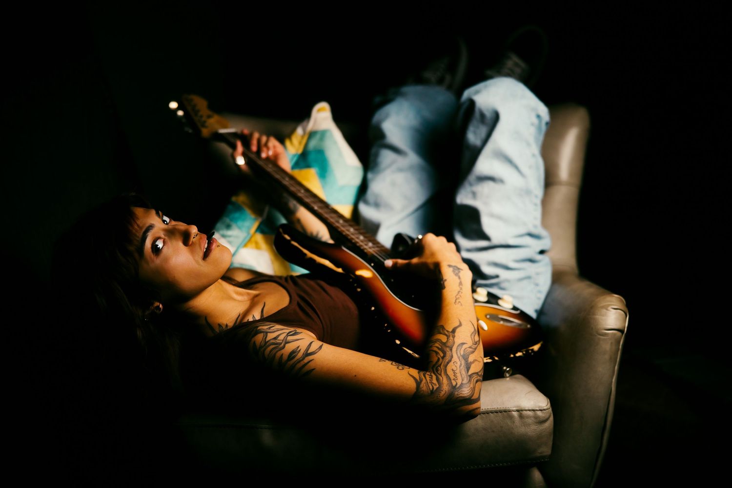 Tattooed woman lying on couch playing guitar in moody dark setting.