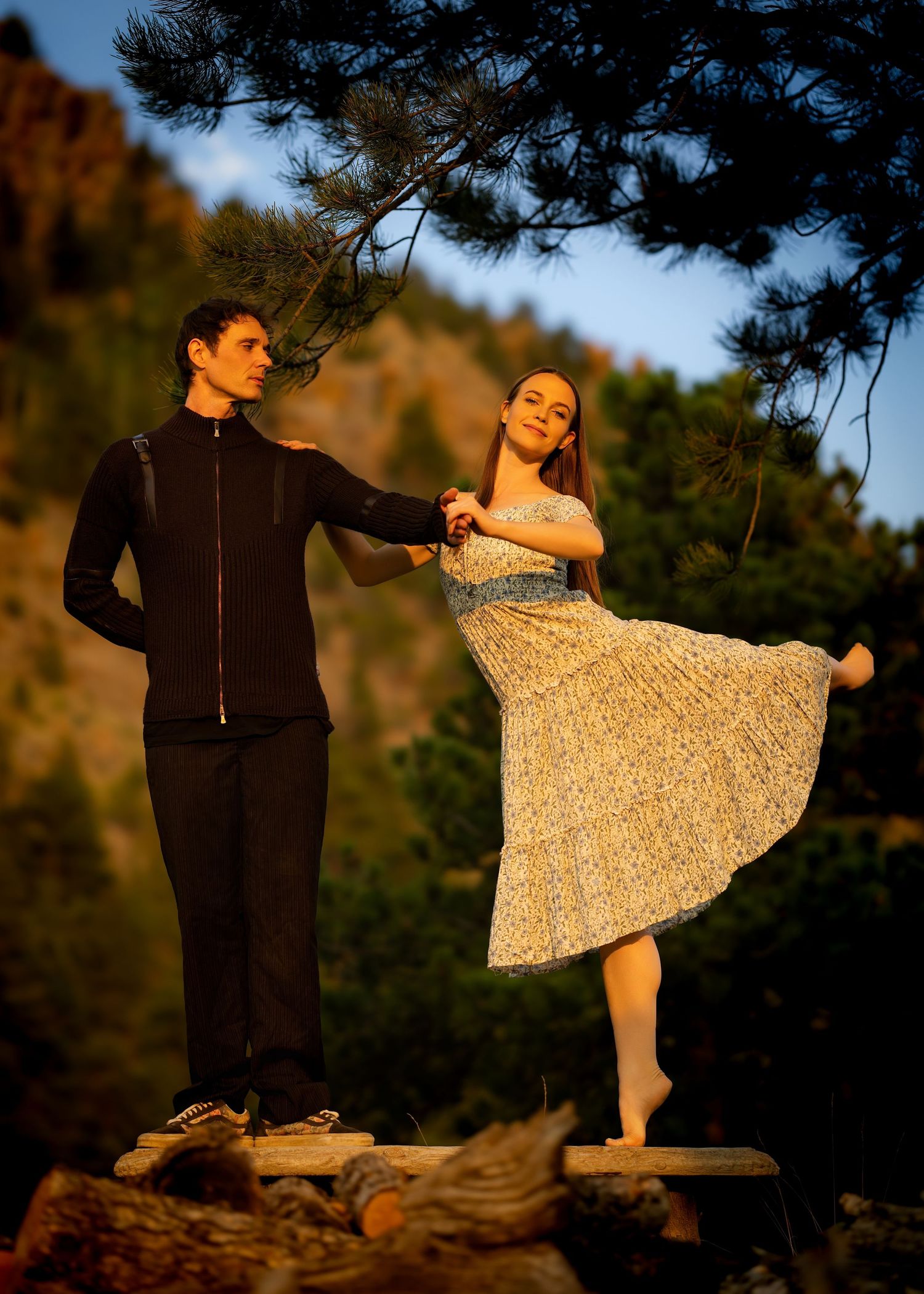 Couple dancing outdoors on wooden beam at golden hour in mountain setting.