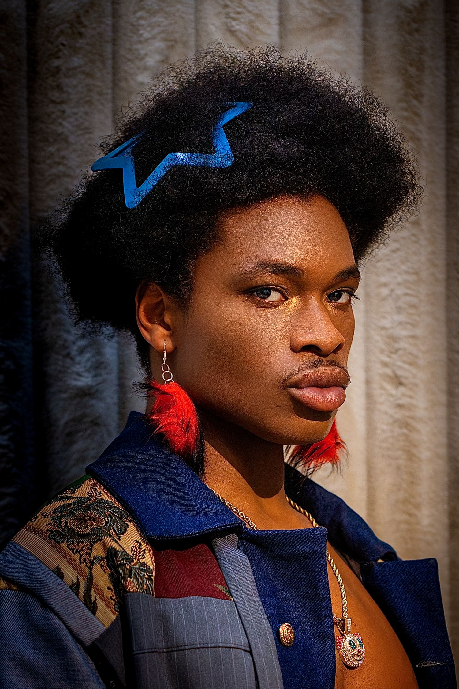 Close-up portrait of male model with afro, blue crown hair clip, and red feather earrings.