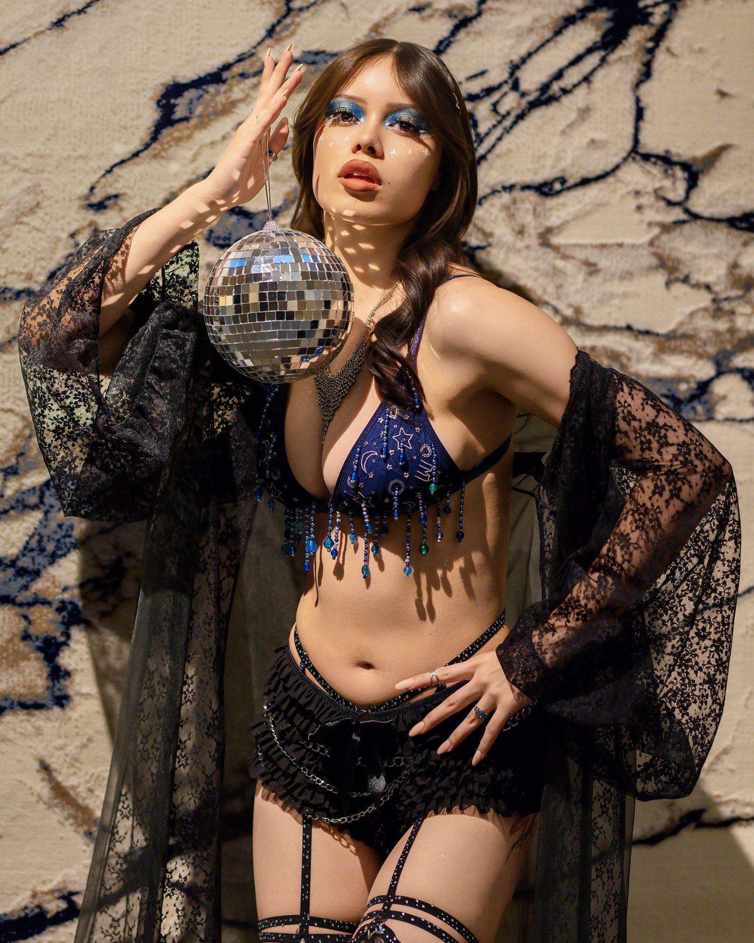 Bold fashion model in sequined blue bikini and lace robe holds disco ball against marble wall.