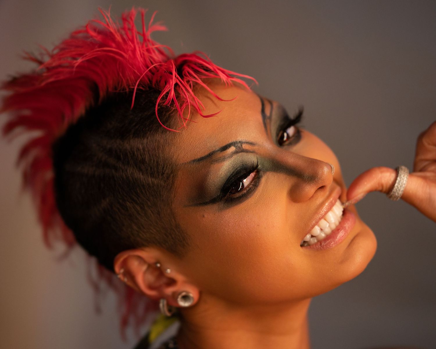 Woman with vibrant red mohawk and bold green eye makeup smiling confidently.