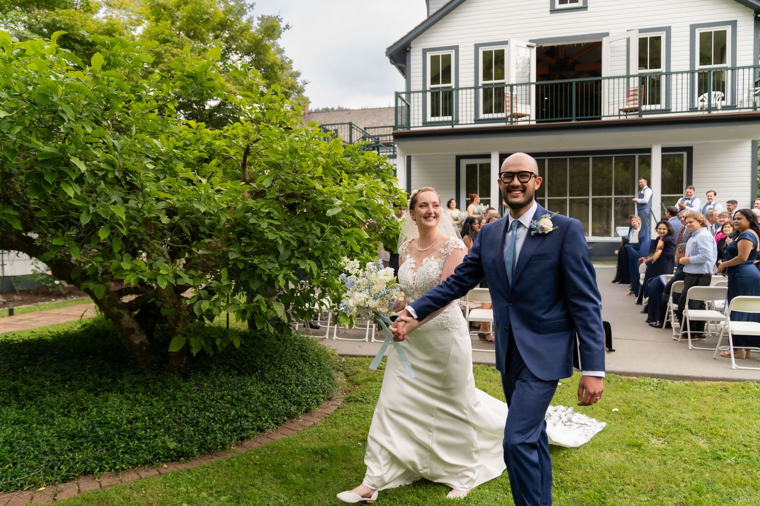 Bride and groom walking hand in hand through lush garden after outdoor wedding ceremony at elegant white venue.