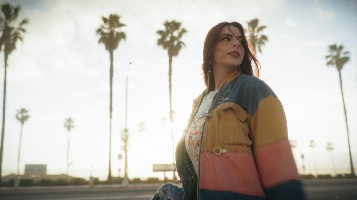 Young woman in colorful striped jacket standing outdoors with tall palm trees and golden sunset sky behind her.