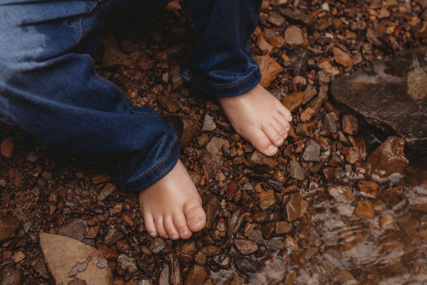 Barefoot child standing on wet rocky ground wearing blue jeans, exploring nature outdoors.