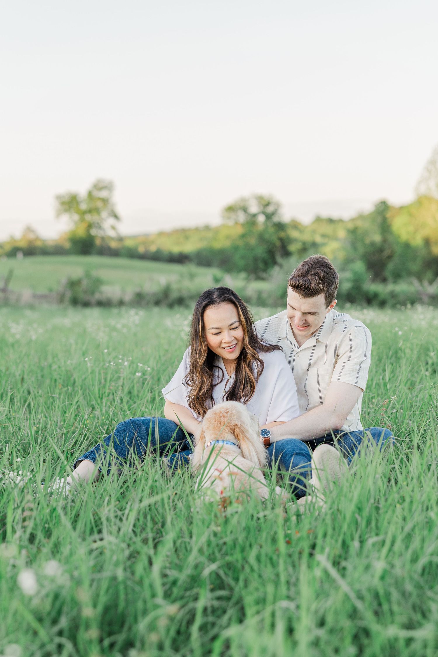 A couple sits together in a lush green field, smiling and petting a small dog on a sunny day.