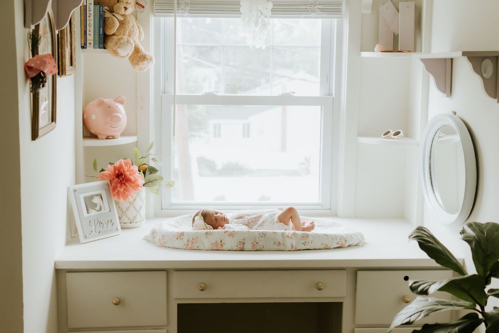 Bright nursery changing table with a newborn baby, floral arrangement, framed photo, and stuffed animals near a sunlit window.