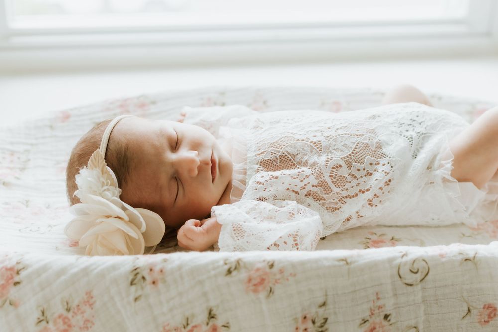 Peaceful newborn baby sleeping in a floral bassinet wearing a delicate white lace outfit and bow headband.