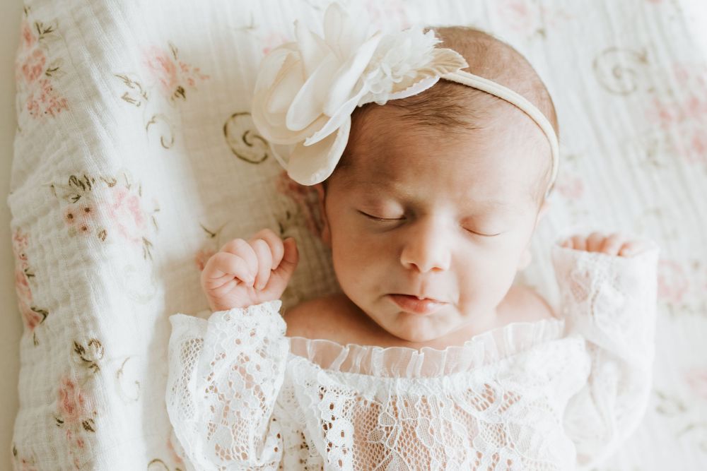 Peaceful newborn baby girl sleeping in white lace outfit and bow headband on floral blanket.