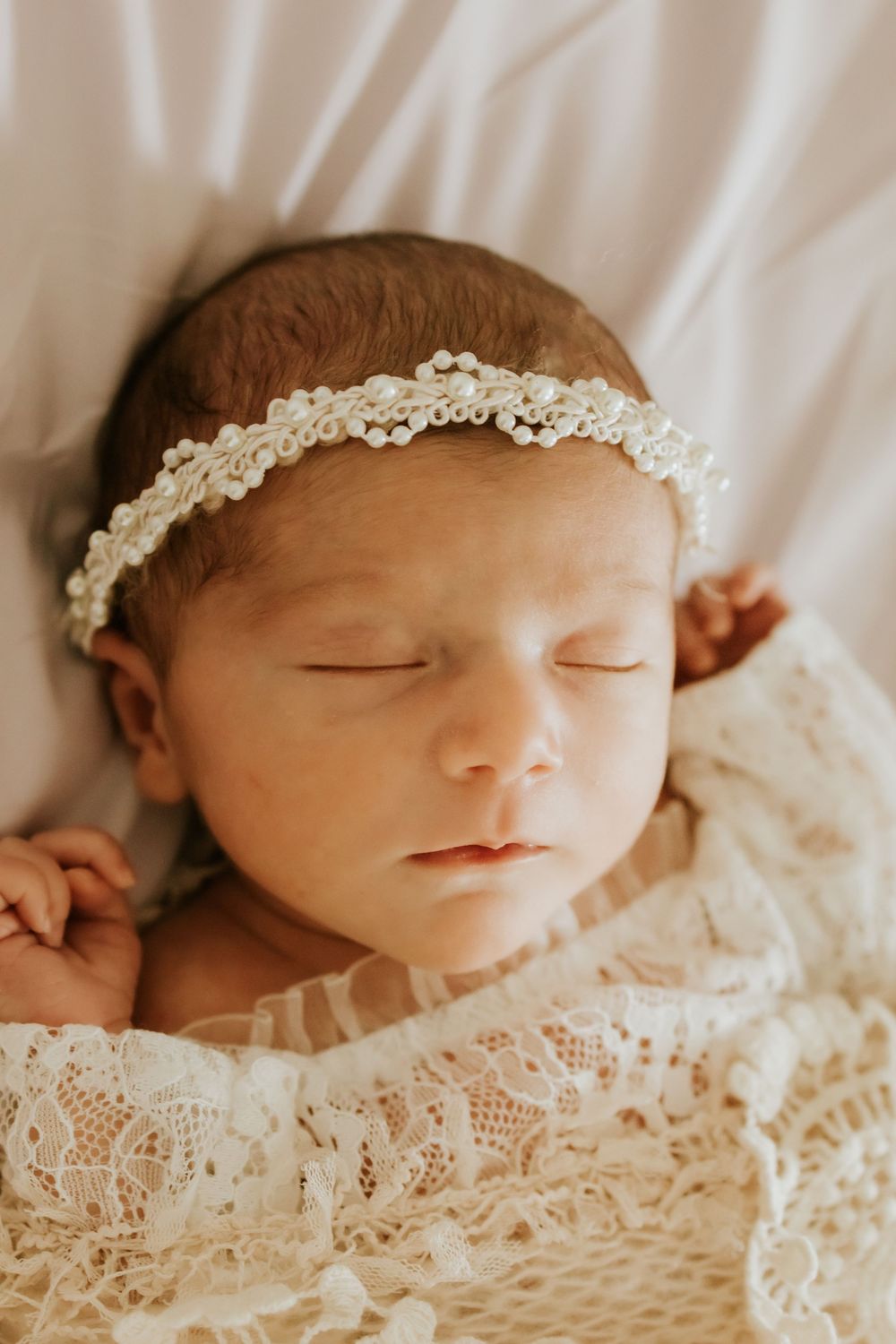 Sleeping newborn baby girl wrapped in cream lace swaddle wearing a delicate pearl and lace headband.