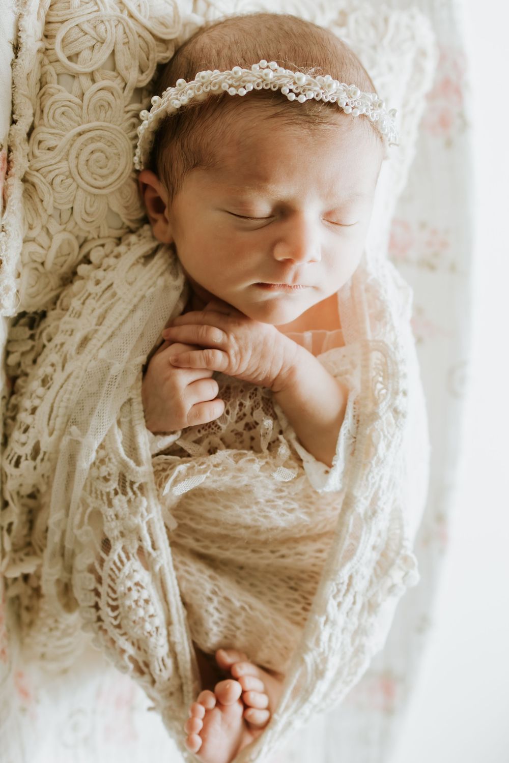 Sleeping newborn baby wrapped in cream lace swaddle with pearl headband in a soft floral bassinet.