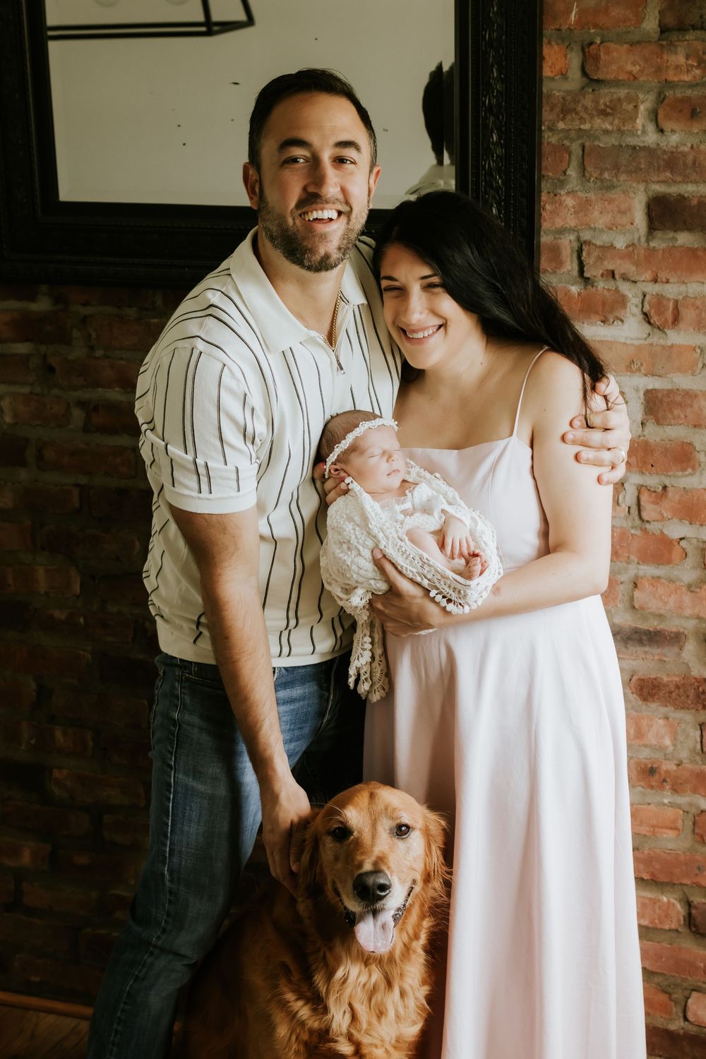 Happy couple smiling and holding their newborn baby girl with a golden retriever dog in front of a brick wall.