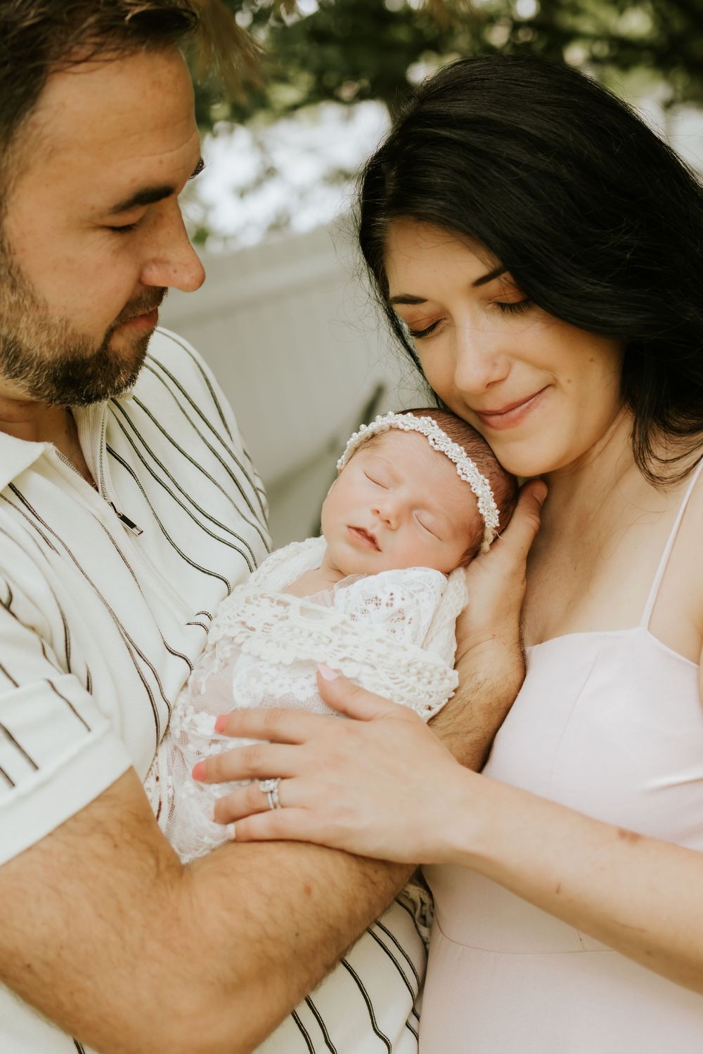 Happy parents holding their sleeping newborn baby wrapped in white lace during an outdoor family photo session.