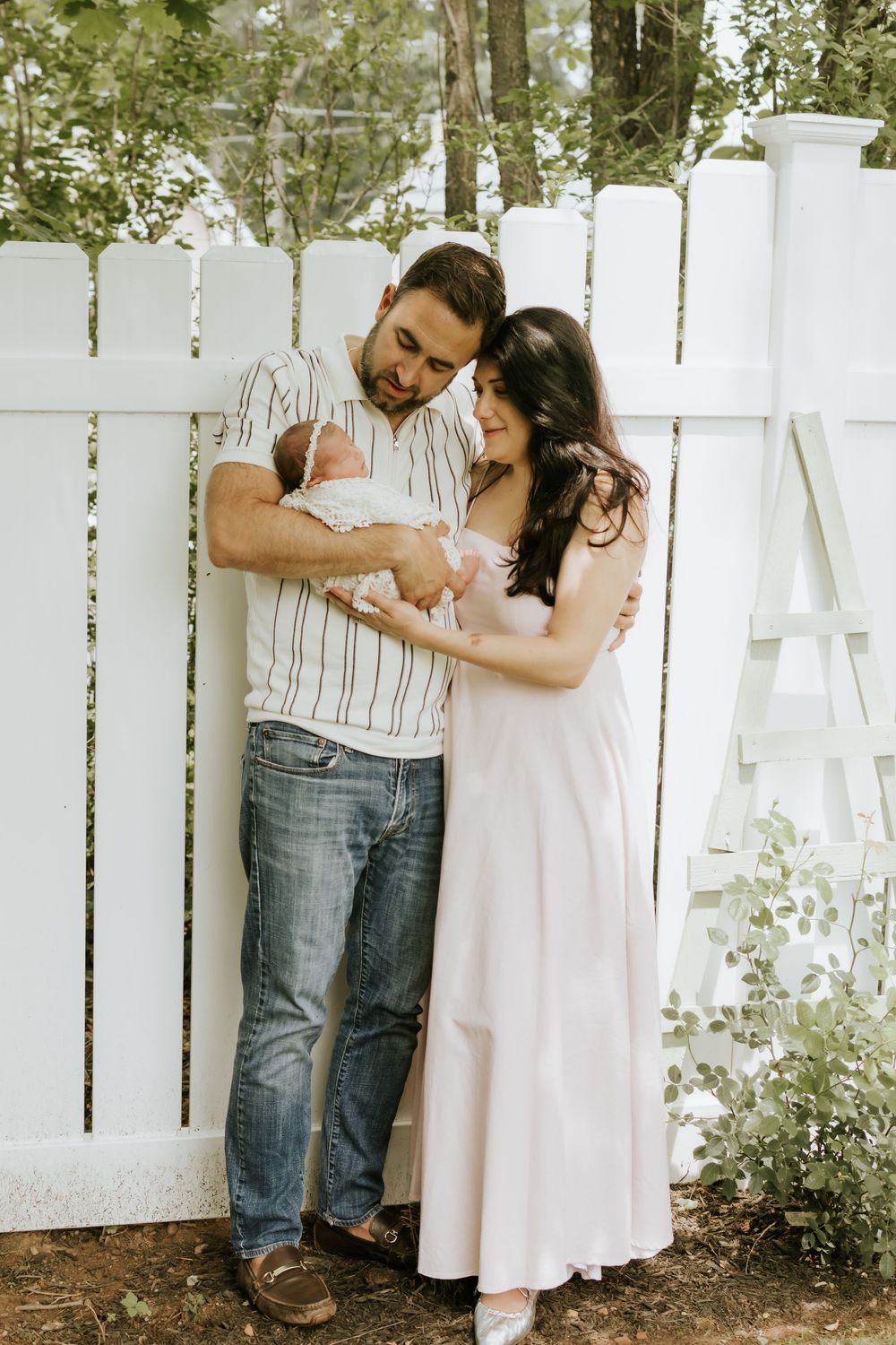 A couple in casual attire lovingly gazes at their newborn baby by a white picket fence outdoors.
