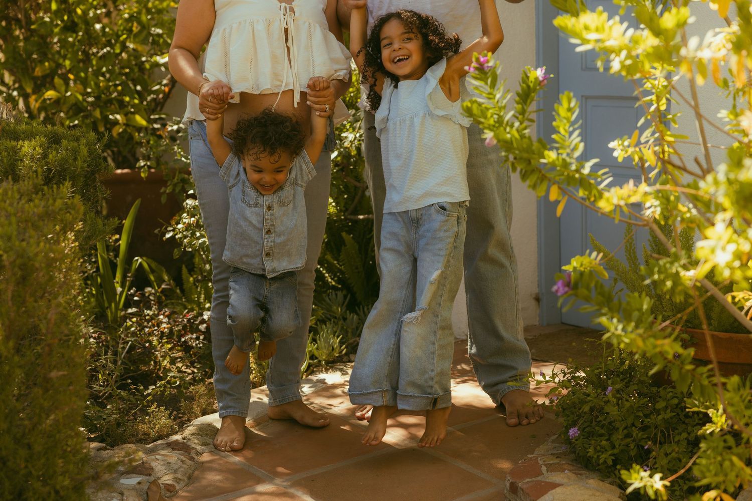 Two children joyfully hold hands with adults while walking barefoot on a garden path surrounded by lush green plants.