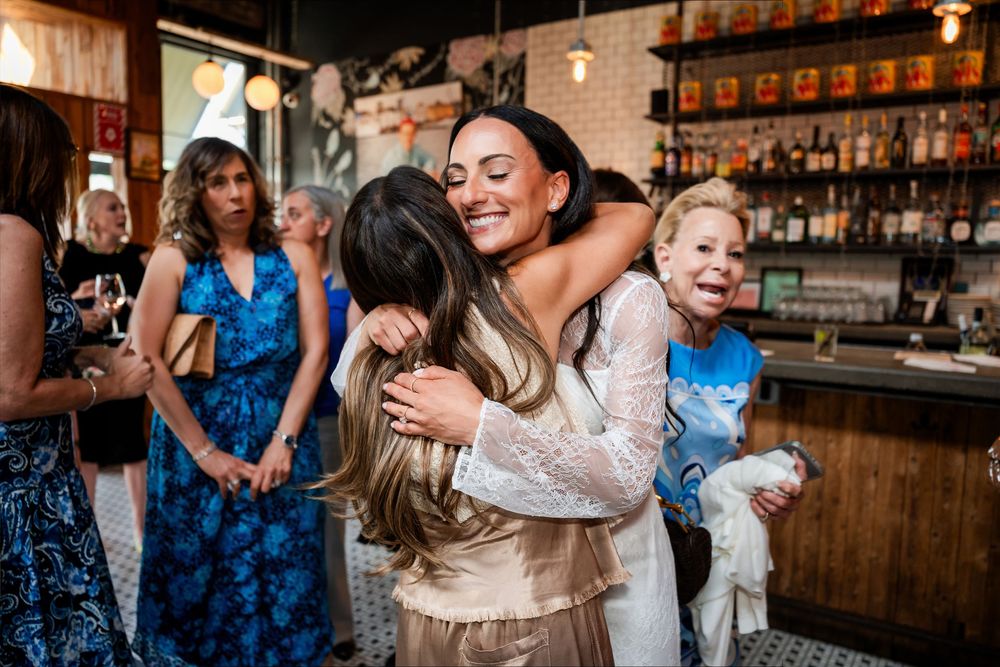Bride in white lace dress shares joyful hug with guest at wedding reception near a bar.