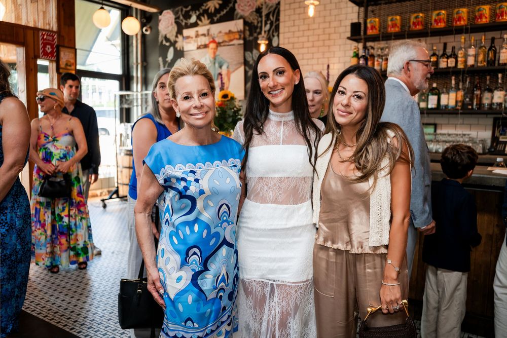Three elegantly dressed women pose together smiling at a stylish restaurant event.