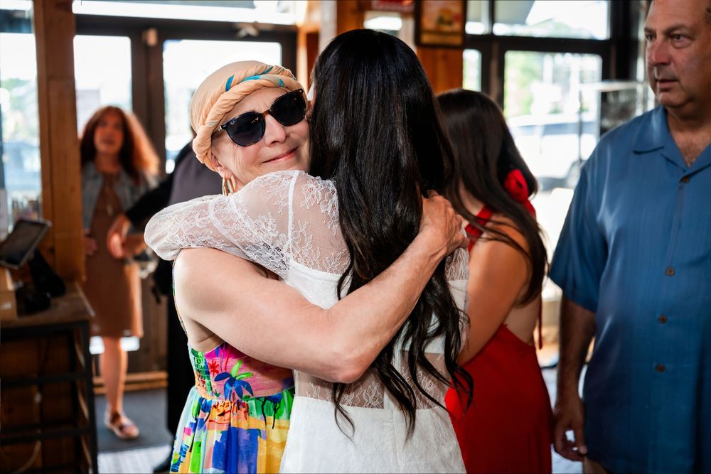 Two women share a warm hug at an indoor gathering, one wearing sunglasses and a colorful dress.