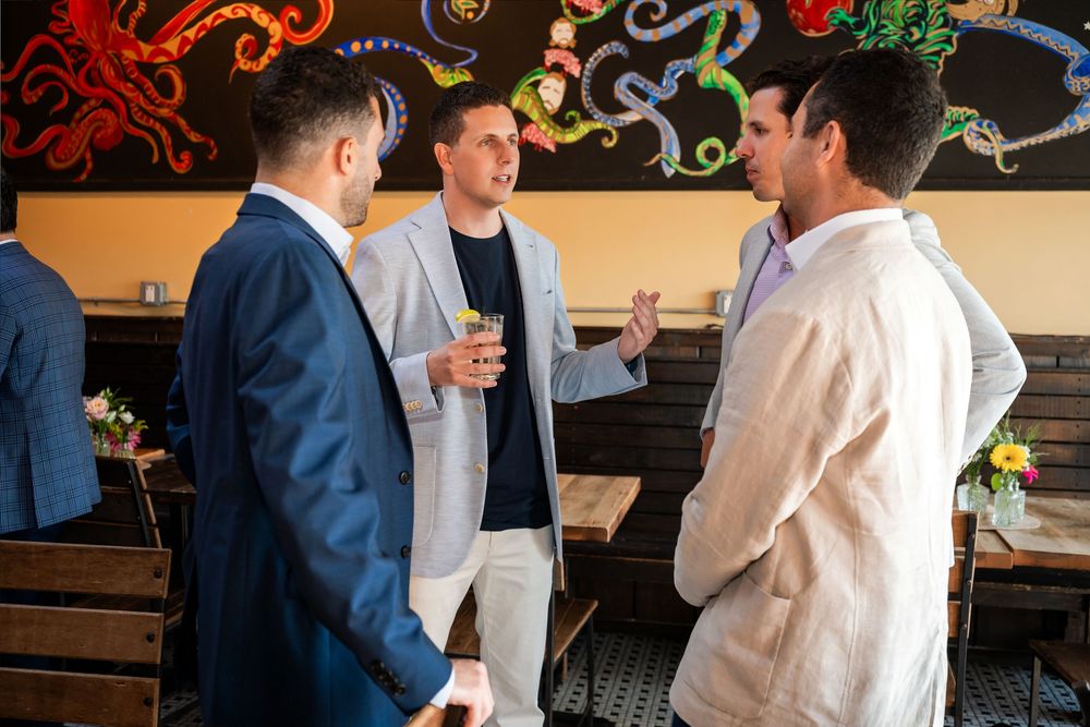 Three men in smart casual attire converse in a stylish restaurant with colorful wall art.