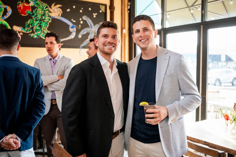 Two smiling men in blazers pose at an indoor social gathering, one holding a drink, with guests mingling in the background.