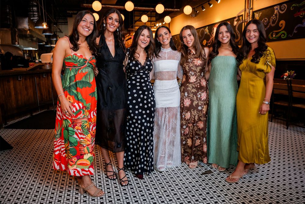 Seven women in colorful dresses posing together and smiling at an indoor restaurant celebration.