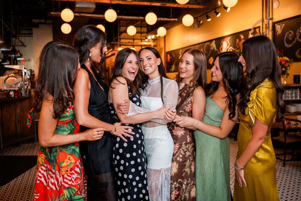 A group of seven women laughing and celebrating together in a warmly lit restaurant, dressed in elegant colorful outfits.