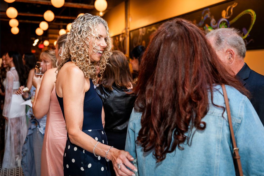 Women with curly hair socializing at an elegant indoor event with warm lighting and decorative backdrop.