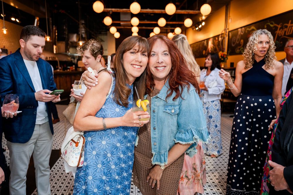 Two women smiling and holding cocktails at a stylish indoor party with warm pendant lights.