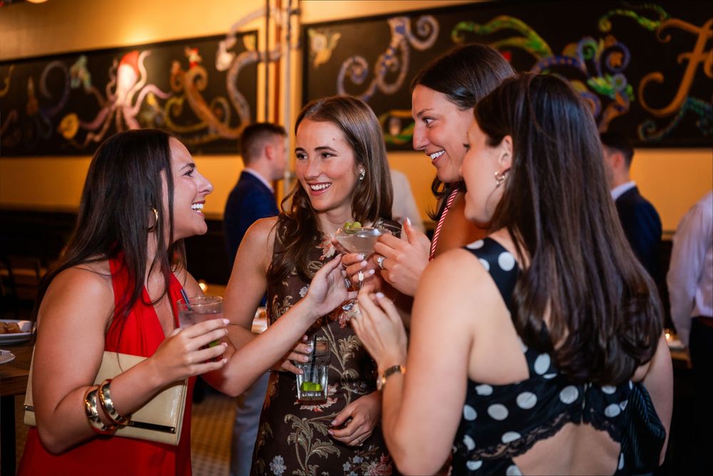 Four women laughing and socializing at an elegant party, holding cocktails in a warmly lit venue.