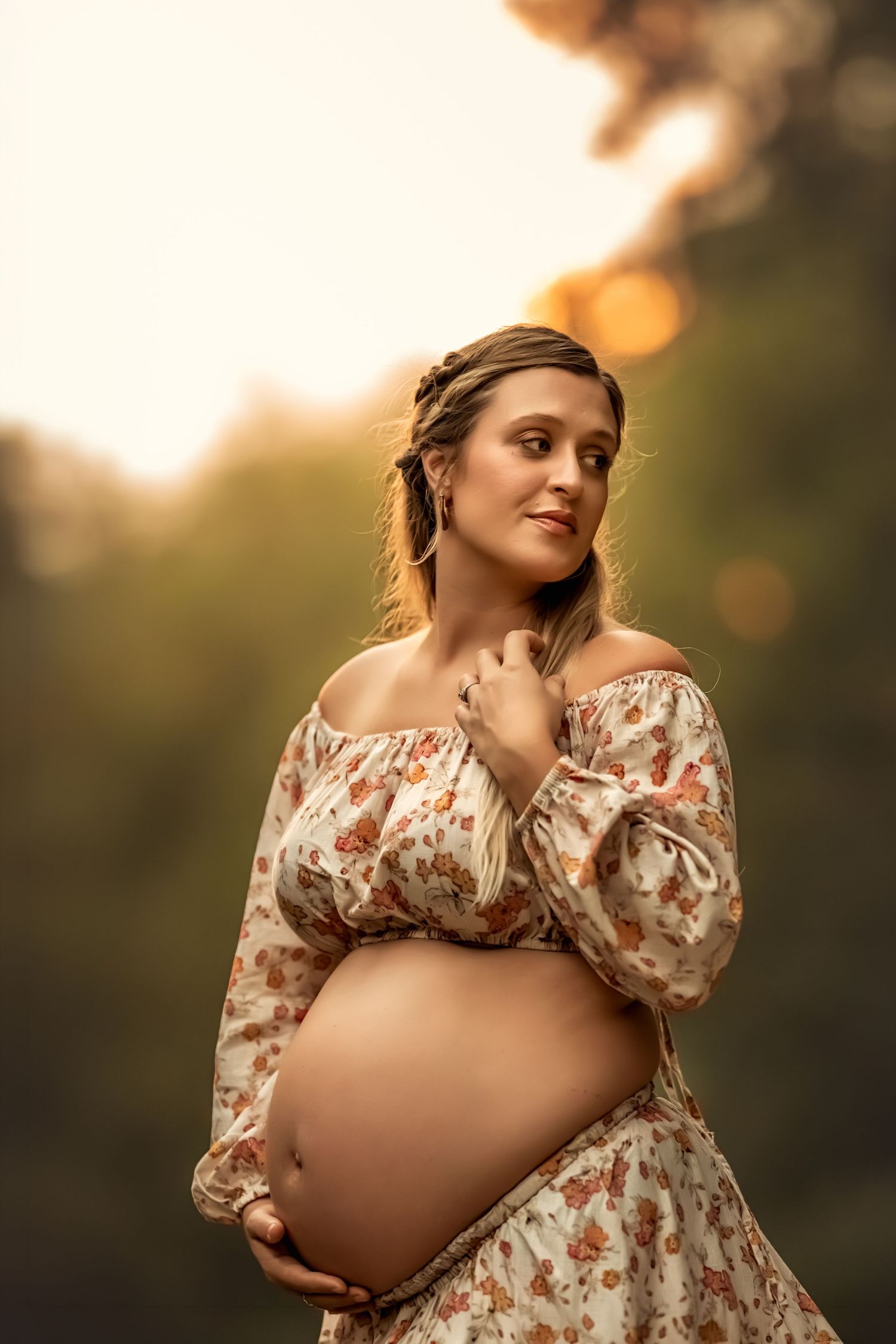 Pregnant woman in floral off-shoulder dress cradling baby bump outdoors in golden sunset light.