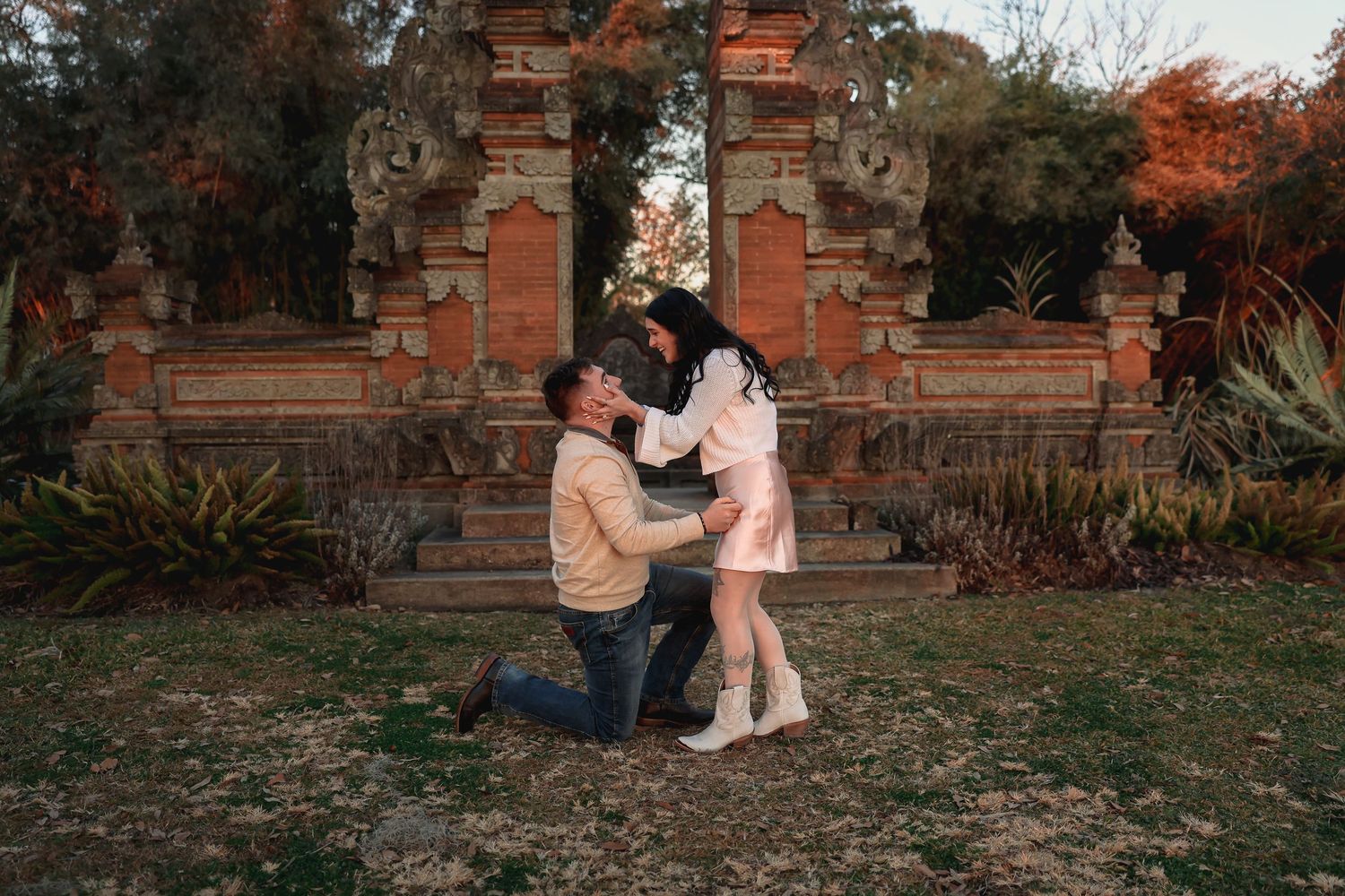 Couple sharing a romantic moment near ornate Balinese stone gates in a lush garden at golden hour.