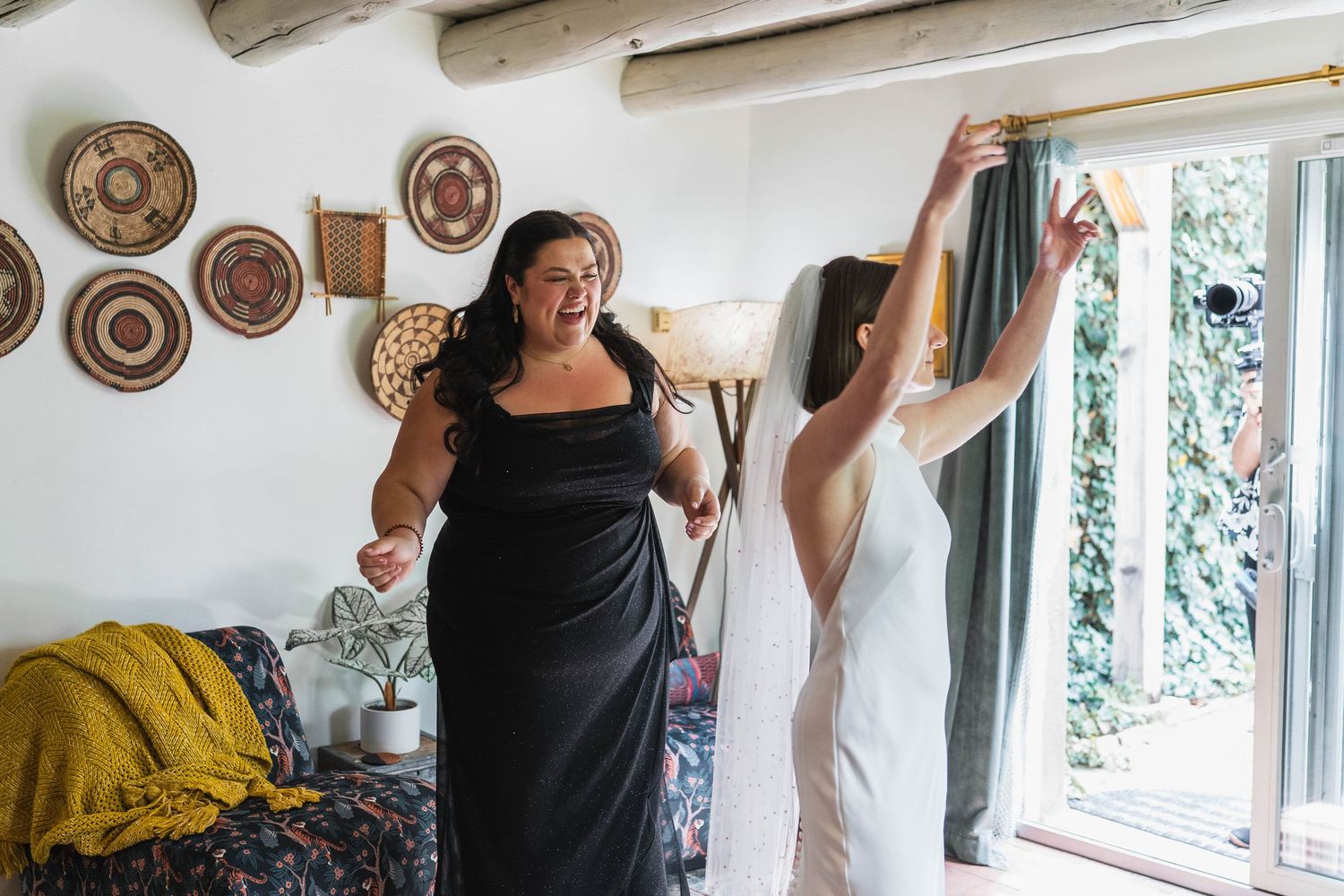Bridesmaid in black gown laughs with bride in white dress adjusting curtains in a rustic decorated room.