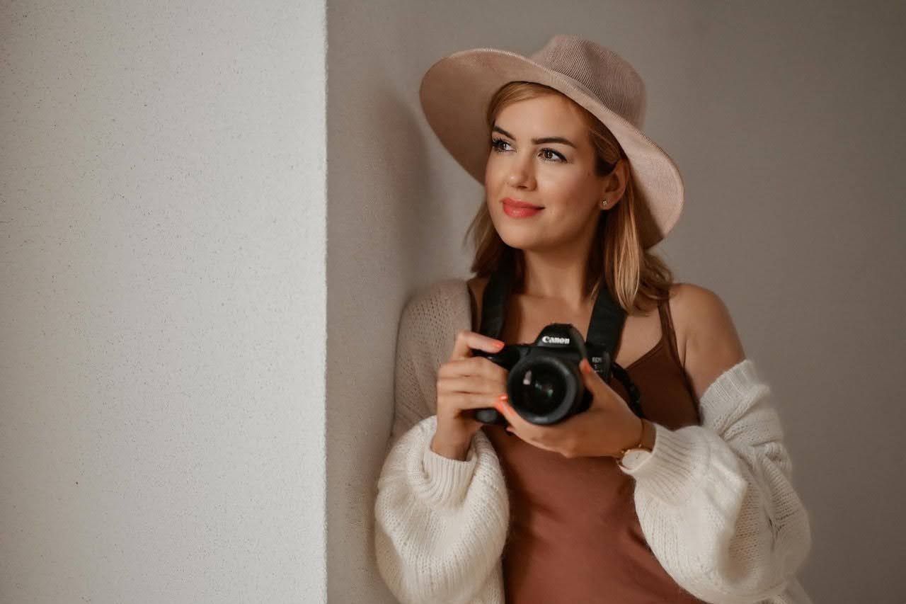 Woman in beige hat holding Canon camera, wearing white cardigan, smiling and looking away in a bright studio setting.