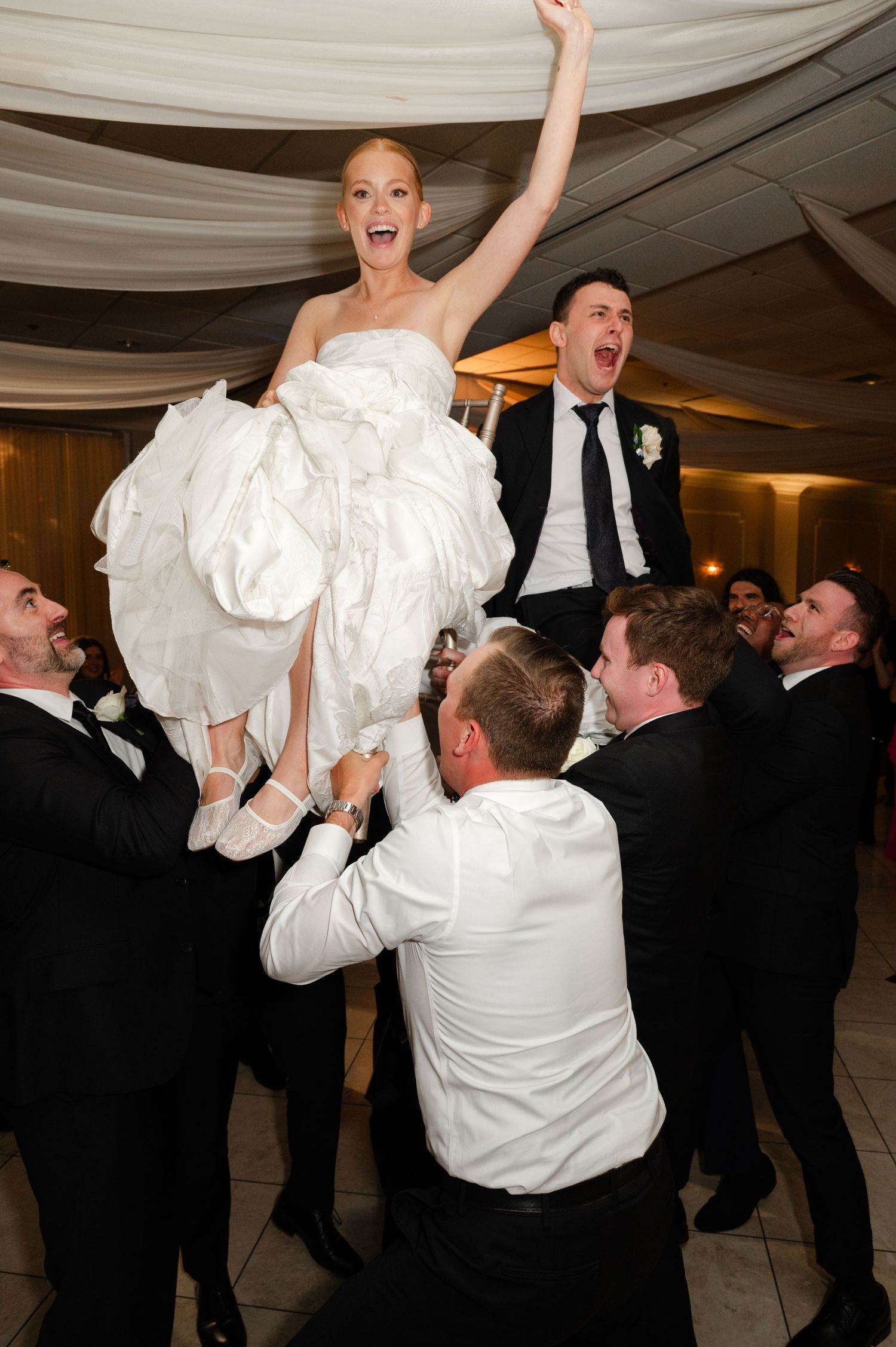 Bride lifted by groomsmen during joyful wedding reception celebration, arms raised in excitement.
