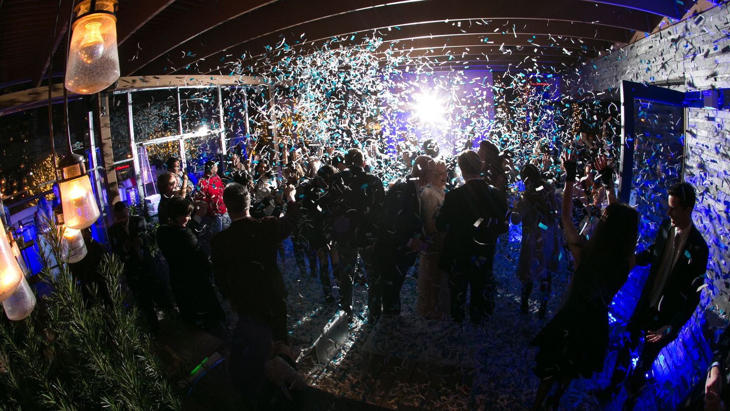 Colorful confetti rains down on a crowd of party guests dancing at a festive nighttime outdoor event with blue lighting.