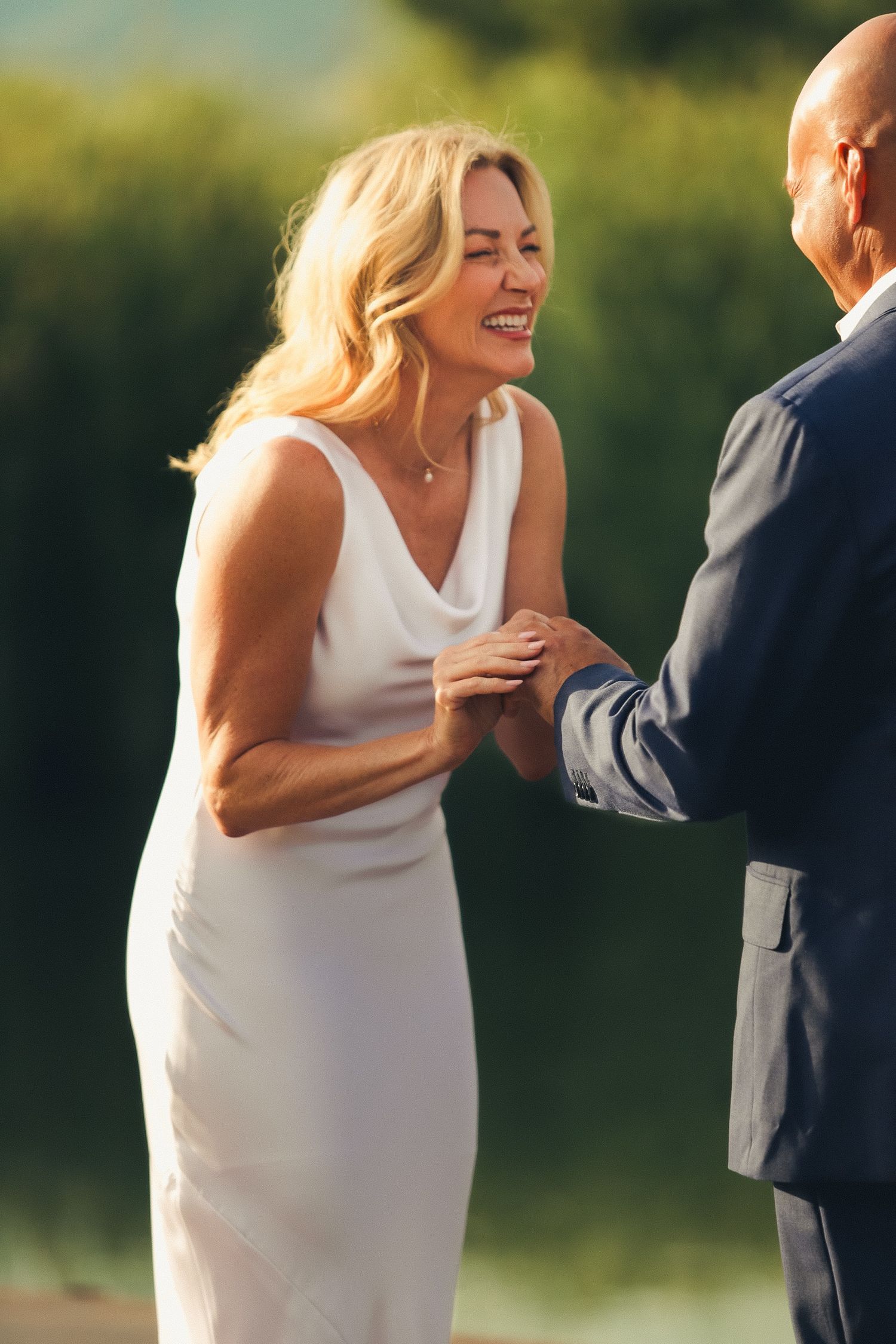 Bride in white dress laughing joyfully while holding hands with groom in navy suit outdoors.