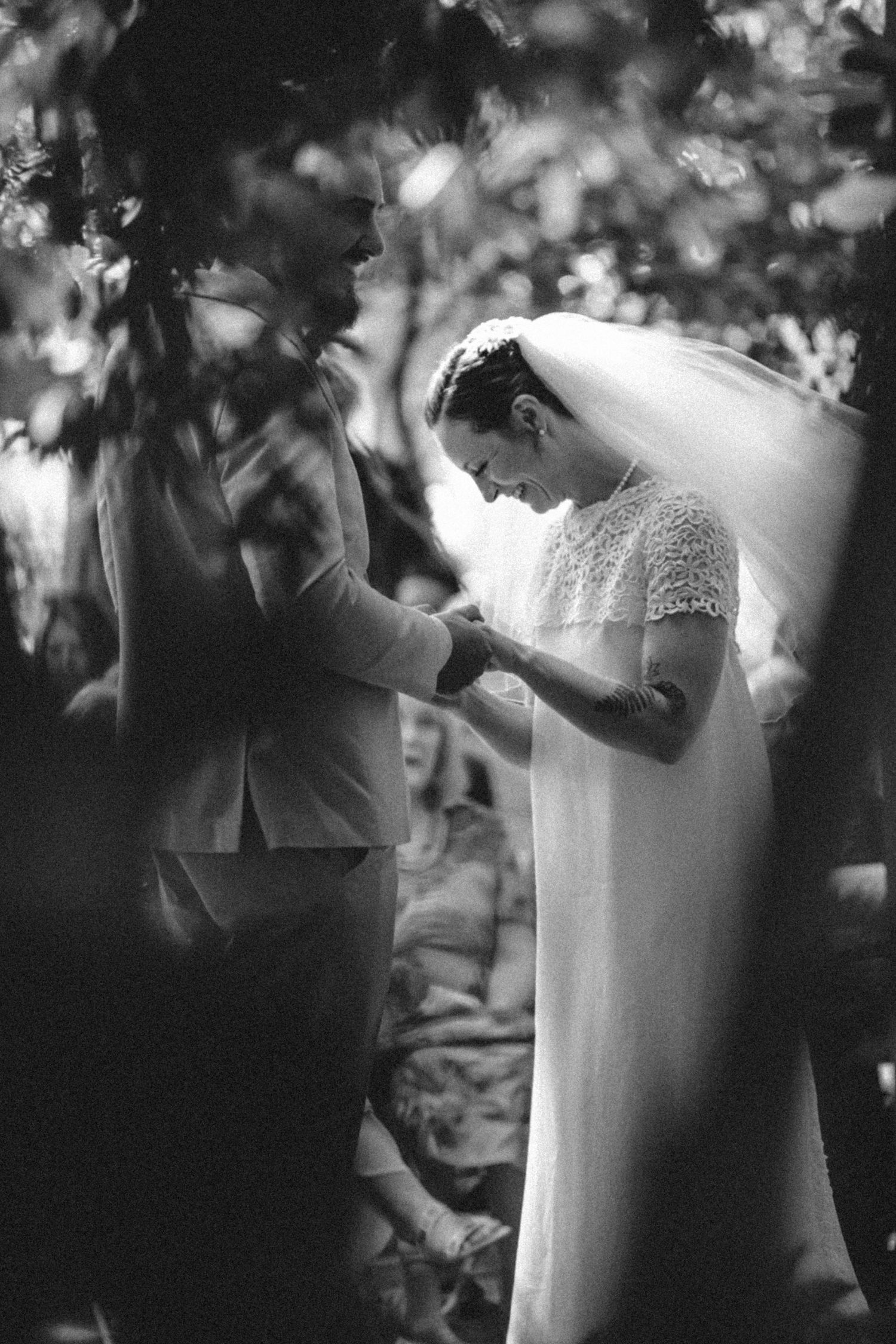 Black and white photo of a bride in a lace veil and groom exchanging rings during an outdoor wedding ceremony.