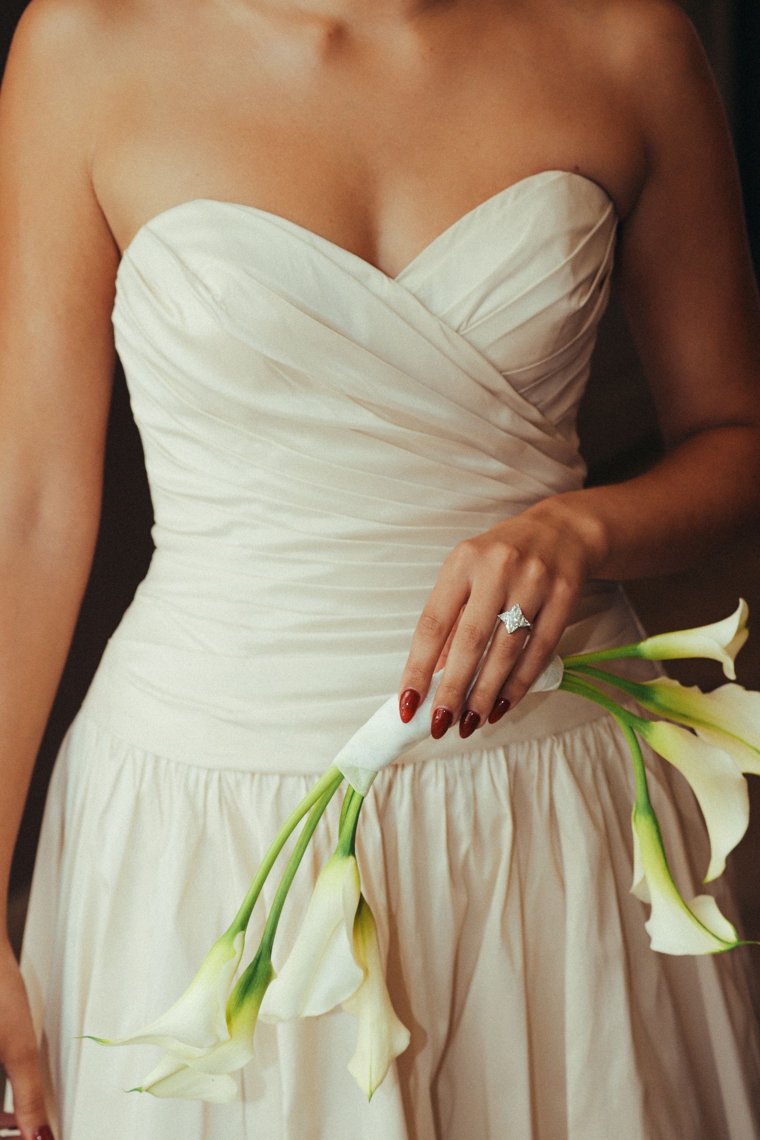 Bride in strapless white gown holding elegant calla lily bouquet, showcasing diamond engagement ring with red manicure.