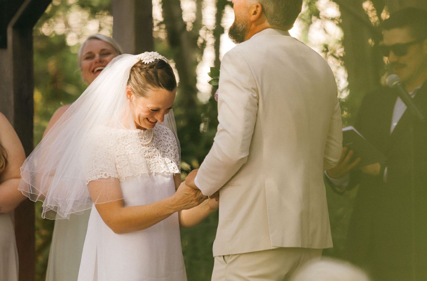 Bride in white dress and veil smiling during outdoor wedding ceremony while holding hands with groom in beige suit.