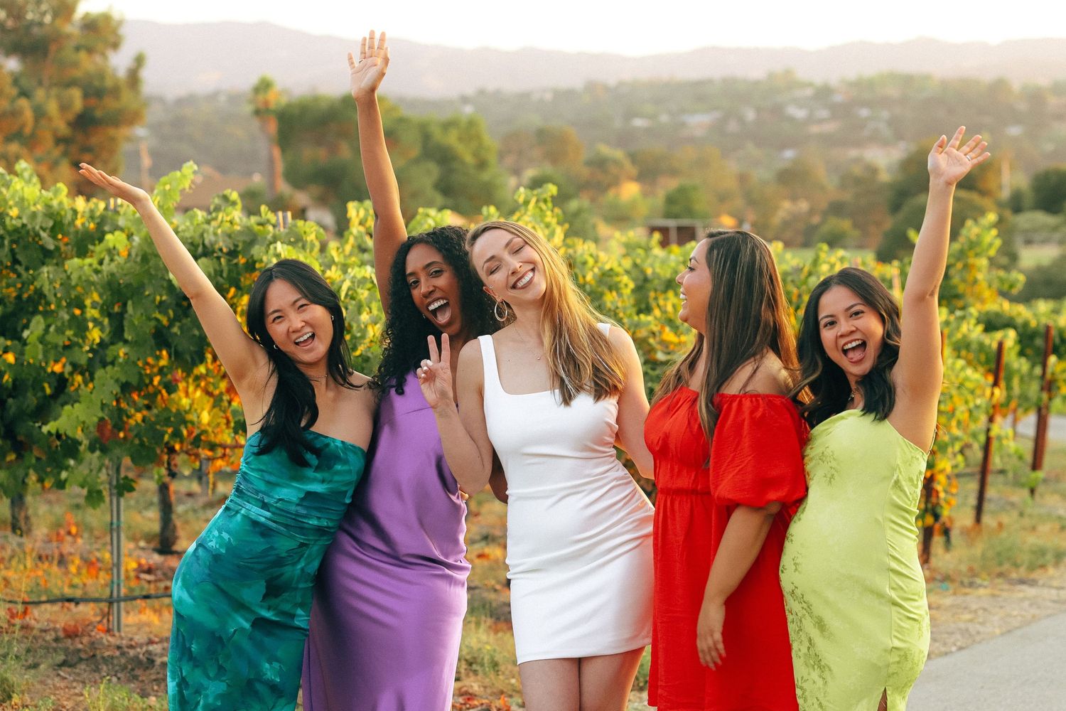 Five women in colorful dresses posing joyfully at a vineyard, raising arms in celebration with lush greenery in the background.