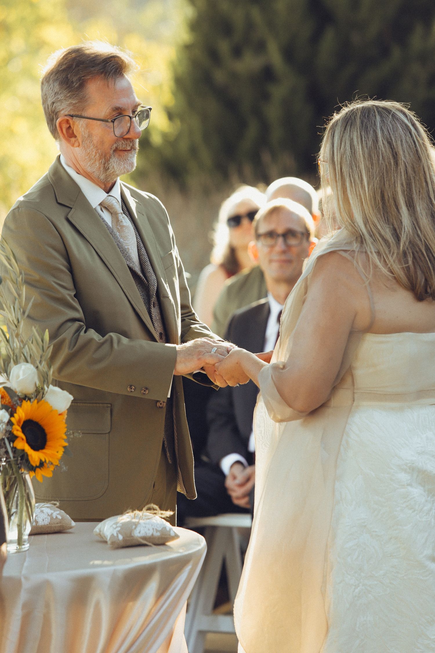 Outdoor wedding ceremony with groom in gray suit holding hands with bride in white dress, sunflowers nearby.