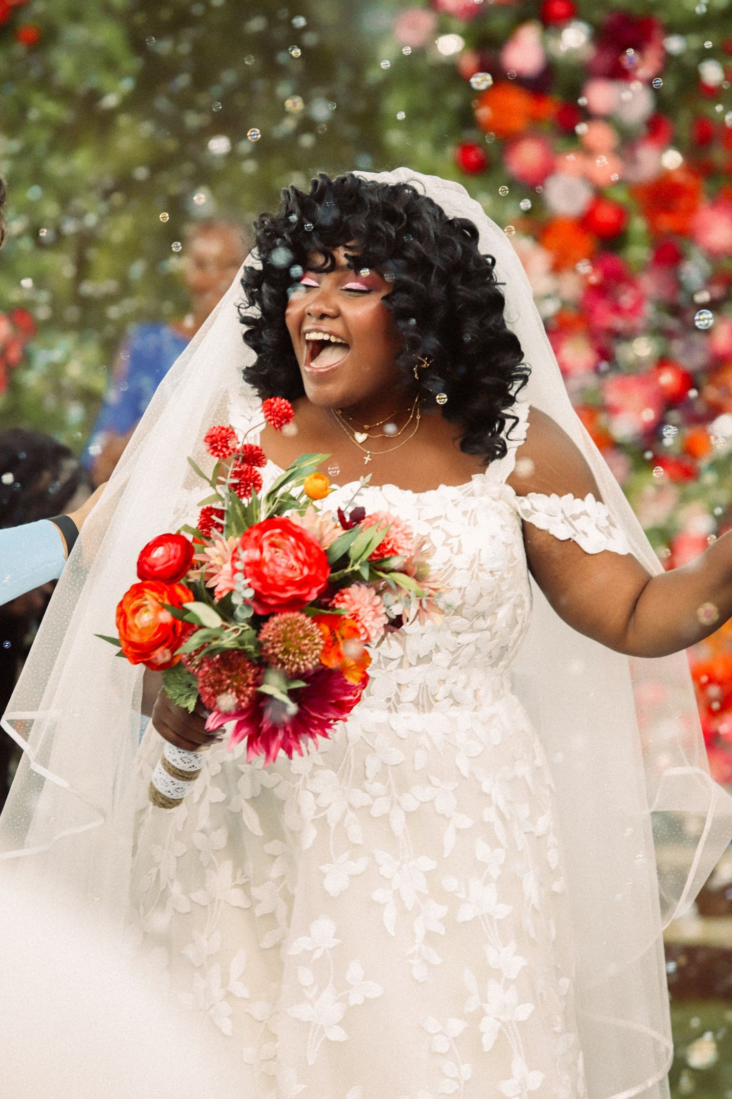 Joyful Black bride in lace gown holding vibrant red bouquet, laughing under confetti at colorful floral wedding ceremony.