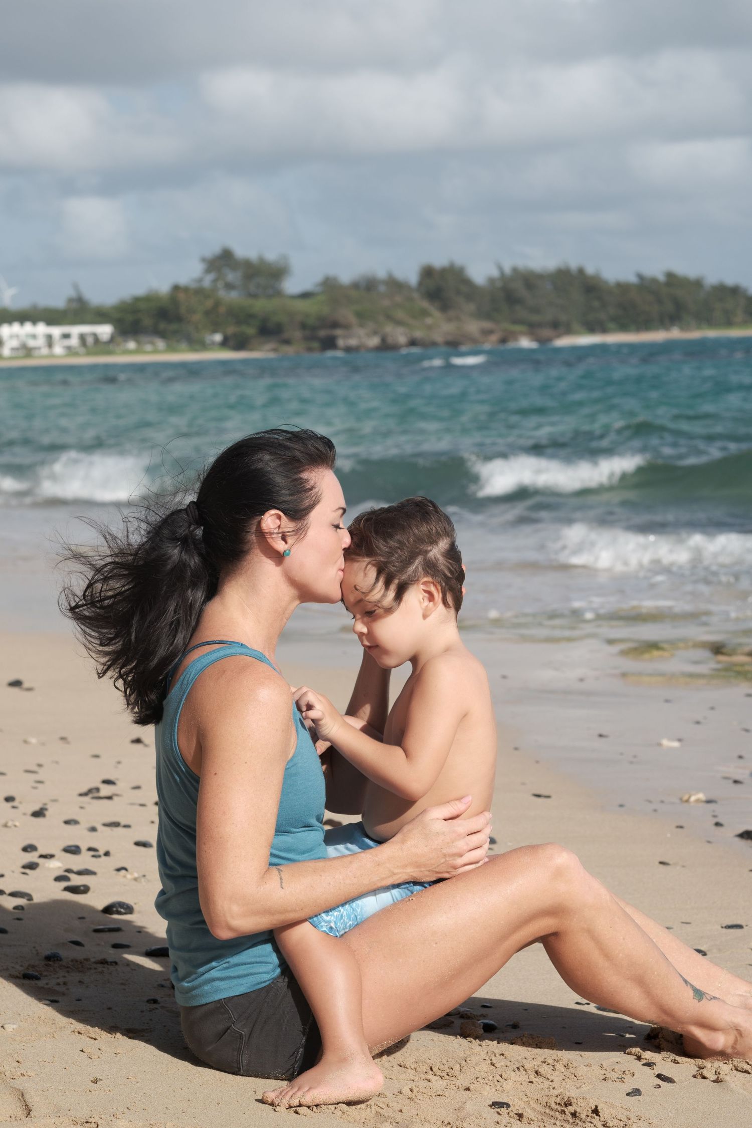 A mother kisses her young child while sitting on a sandy beach with ocean waves and green coastline in the background.