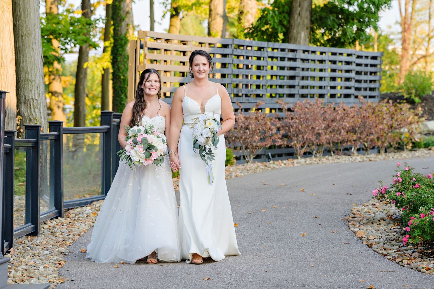 Two brides wearing long white wedding gowns walking down path near waterfall at Mapleside Farm after their wedding ceremony in Ohio