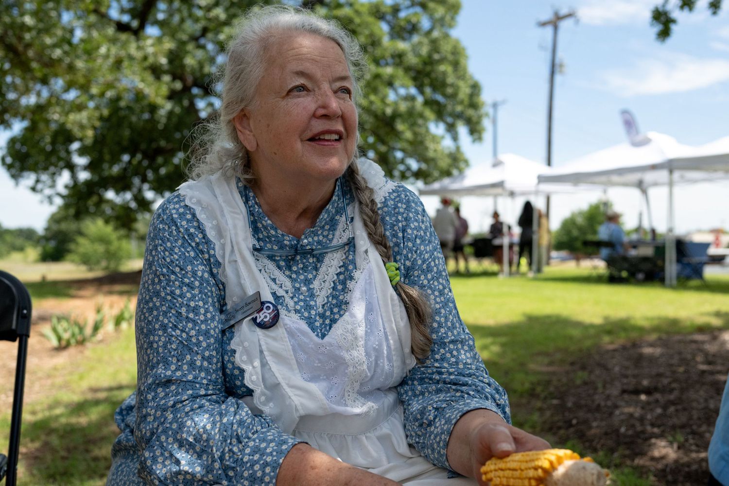 Smiling elderly woman in blue floral dress and white apron holding corn at an outdoor farmers market event.