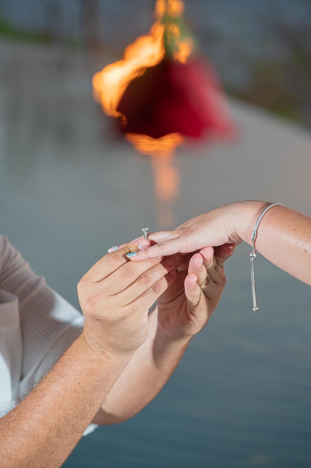 Person placing a diamond engagement ring on a woman's finger near water with a glowing red rose in the background.