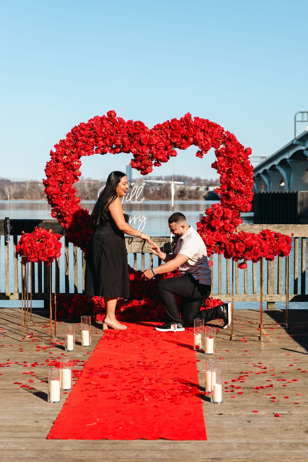 Man proposes to woman on red carpet surrounded by red rose heart arch and candles on a waterfront dock.